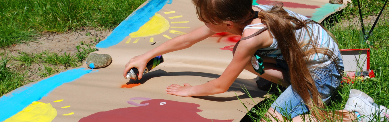 Mała dziewczynka klęcząc maluje farbami na bardzo dużym papierze. Papier leży na trawie.
