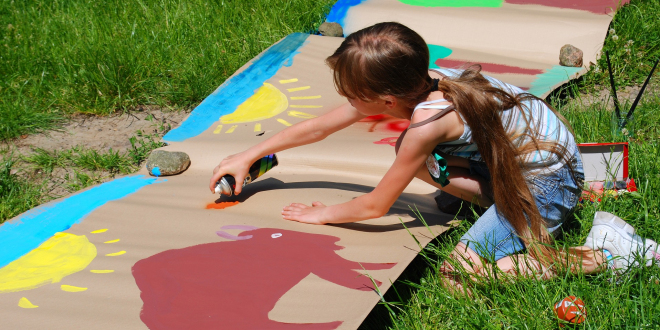 Mała dziewczynka klęcząc maluje farbami na bardzo dużym papierze. Papier leży na trawie.