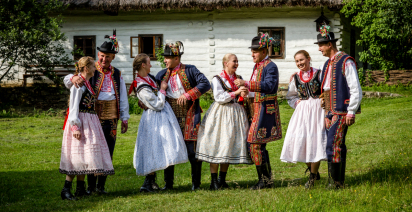 ludzie ubrani w regionalne stroje stojący na trawie przed tradycyjną chałupą