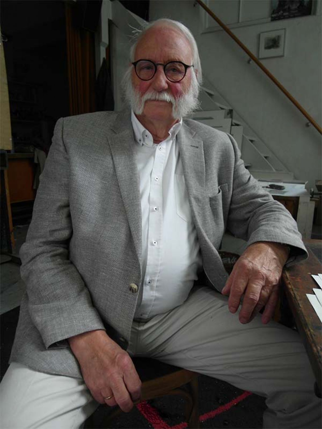 Portret Jana Wagnera. Starszy mężczyzna z siwymi włosami oraz brodą. Ubrany w szarą marynarkę i spodnie oraz białą koszulę. Siedzi na krześle w swoim mieszkaniu.