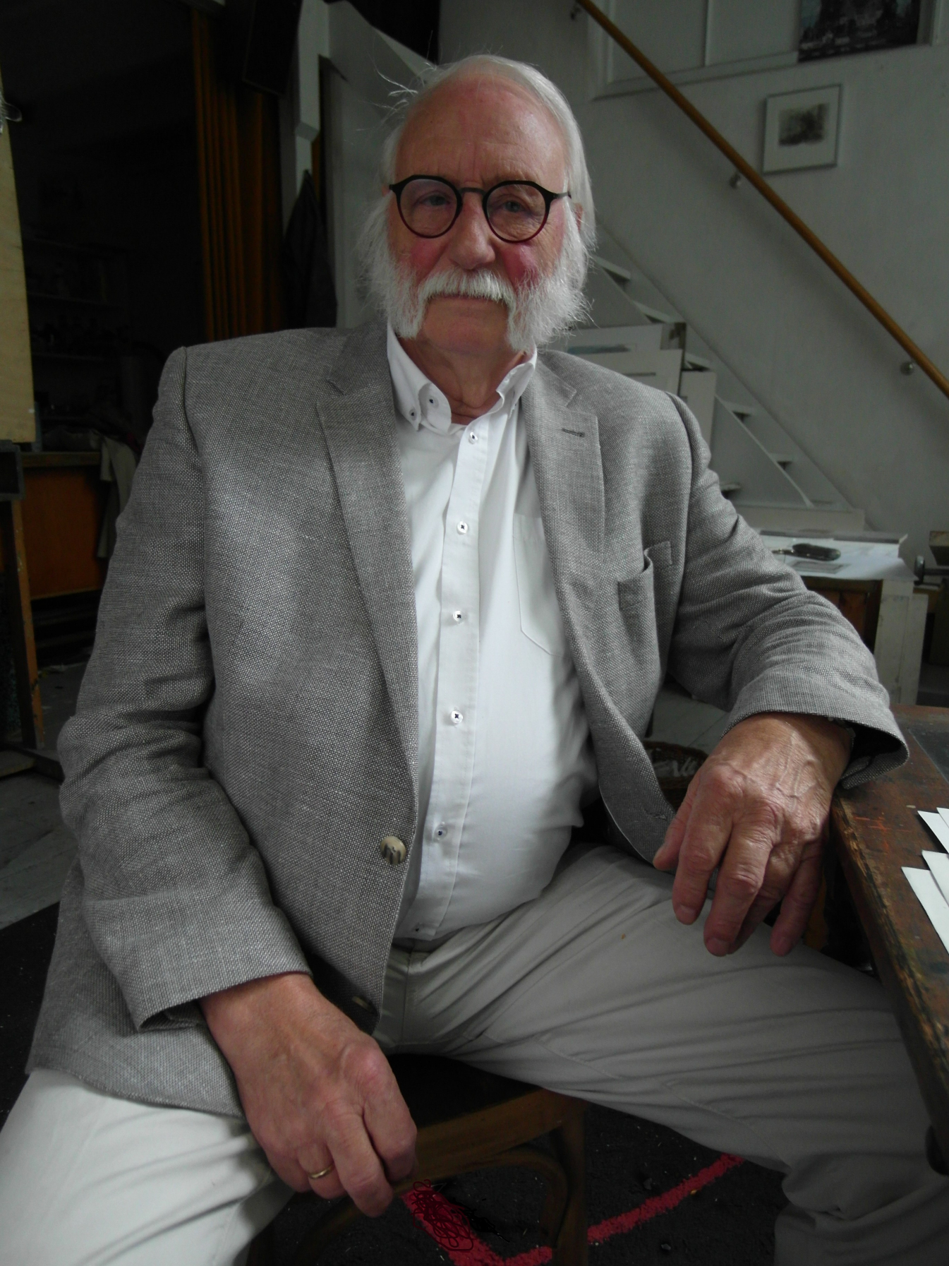 Portret Jana Wagnera. Starszy mężczyzna z siwymi włosami oraz brodą. Ubrany w szarą marynarkę i spodnie oraz białą koszulę. Siedzi na krześle w swoim mieszkaniu.
