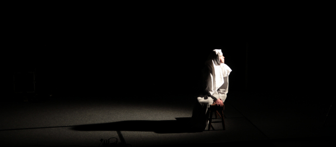 Osoba ubrana w białe szaty siedzi na krześle w świetle reflektora