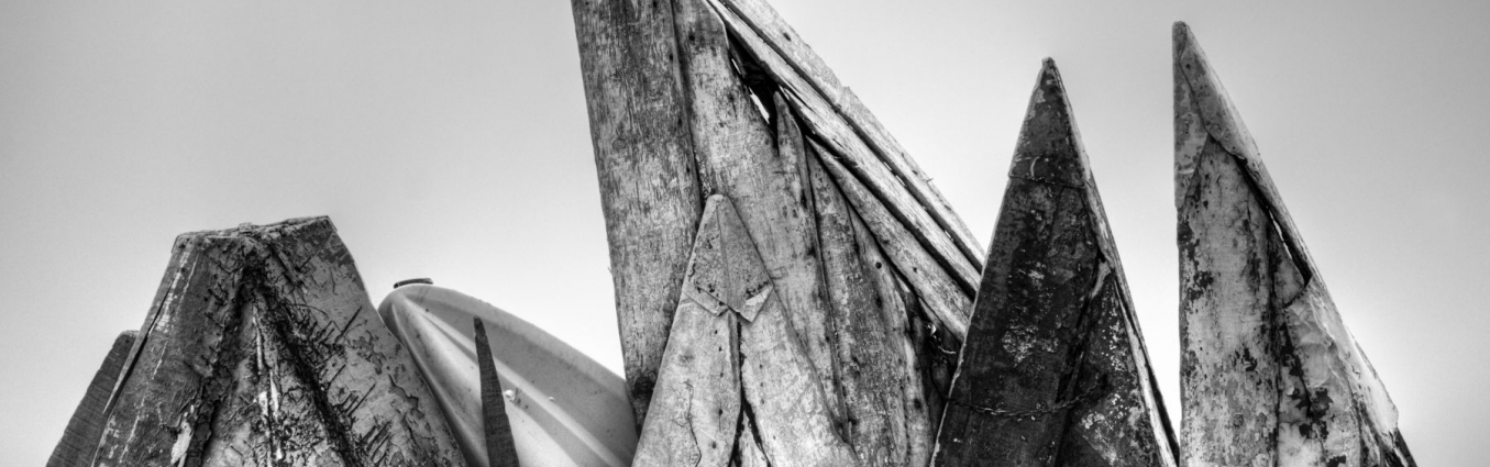 Czarno-białe zdjęcie. Drewniane łodzie postawione pionowo, kompozycyjnie tworzą układ przypominający góry. Drewno jest stare, spękane i dziurawe.