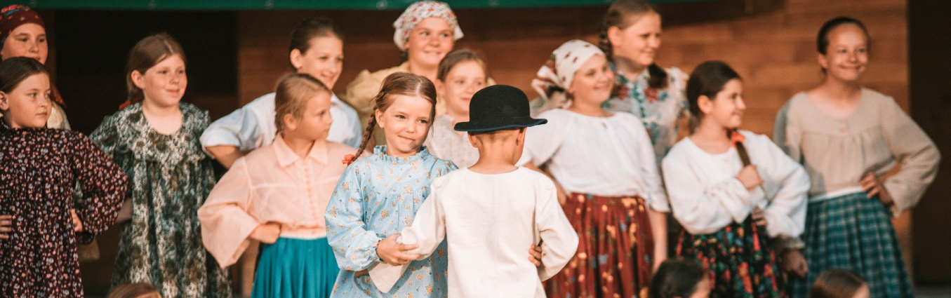 Dziewczynka i chłopiec w strojach regionalnych tańczą na scenie. W tle stoi grupa dzieci również w strojach regionalnych