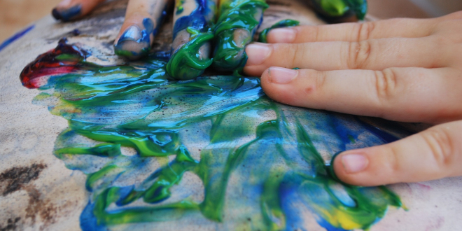 Dziecięce dłonie zatopione w niebieskiej i zielonej farbie. Dziecko wykonuje nieokreślony wzór na papierze. Papier jest już mokry od farby, zaczyna lekko falować.