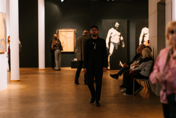 Sala wystawiennicza Galerii Sztuki Współczesnej. Kilka osób spaceruje po sali i ogląda obrazy znajdujące się na ścianach, a kilka siedzi na krzesłach.
