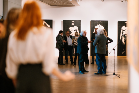Sala wystawiennicza Galerii Sztuki Współczesnej. Na ścianach wiszą obrazy. Na środku w grupie stoją ludzie.