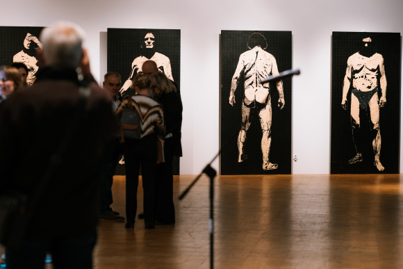 Sala wystawiennicza Galerii Sztuki Współczesnej. Pod białą ścianą ustawione są cztery duże obrazy przedstawiającego postać mężczyzny na czarnym tle. Po lewej stronie grupa ludzi a po prawej statyw z mikrofonem.