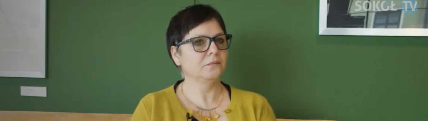 Kobieta w krótkich włosach i okularach siedzi na sofie. W tle zielona ściana.