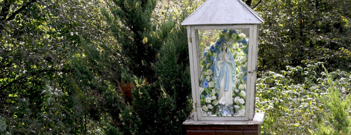 Kapliczka z figurą Maryi. W tle zielone krzewy.